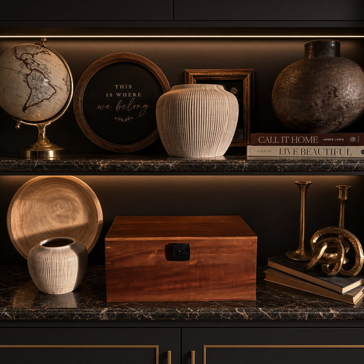 A styled interior shelf display with dark wood cabinetry, marble-look shelves, and warm lighting. Visible objects include a globe, framed wall art, a ribbed cream vase, a textured brown ceramic vessel, books, an acacia Bzz Box stash box, a plate, a small ceramic pot, and decorative brass-toned candlesticks.