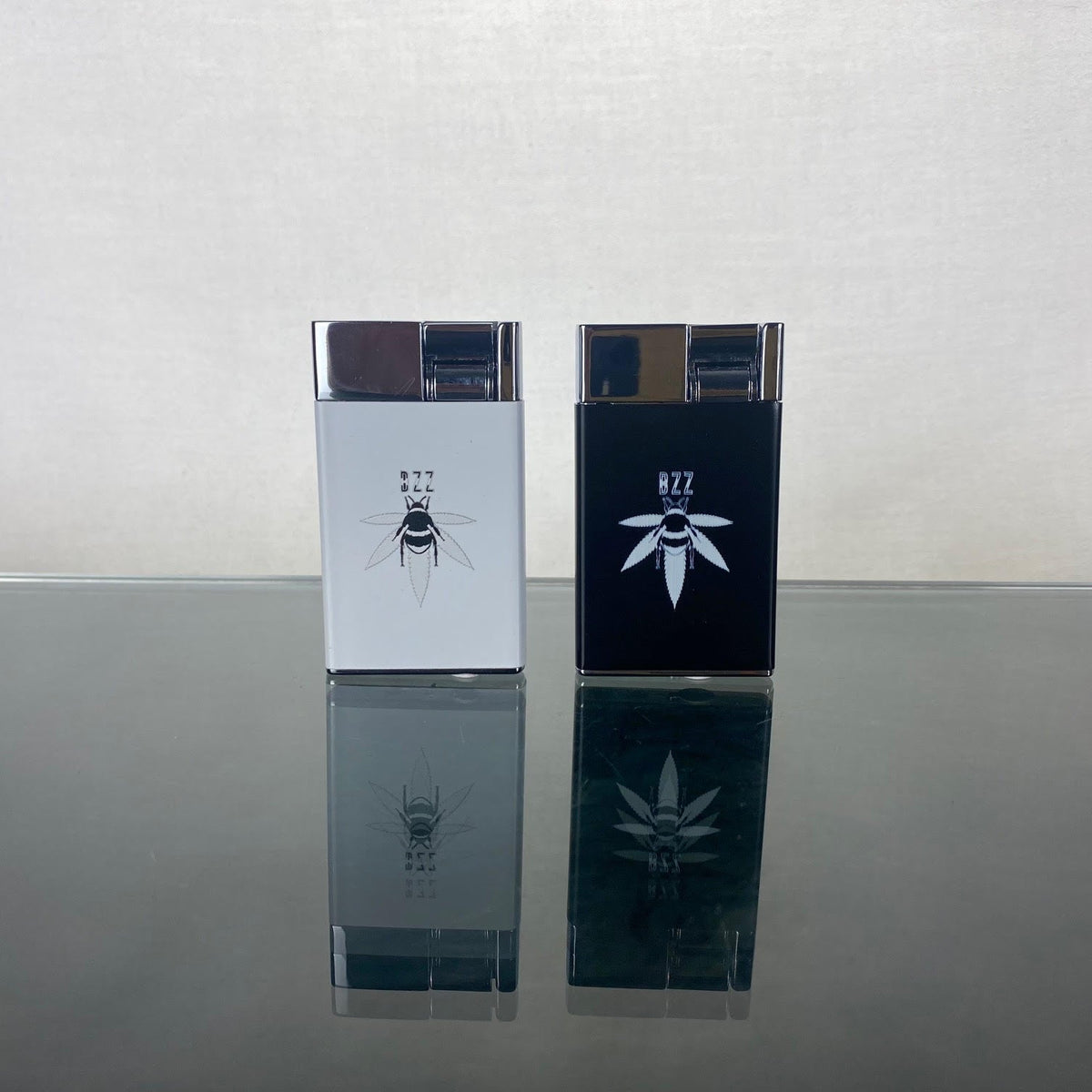 Two rectangular refillable butane lighters with bee designs. The lighters sit on a reflective surface with a light gray background