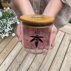 A woman holds a clear glass stash jar with a bamboo lid with gold hygrometer in her hands. The glass is branded with a black Bzz Box logo.