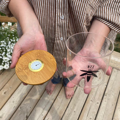 A woman holds a clear glass stash jar in one hand and the bamboo lid with gold hygrometer in her other hand. The glass is branded with a black Bzz Box logo.