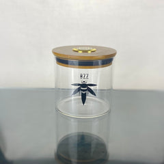 A clear glass stash jar with a bamboo lid with gold hygrometer. The glass is branded with a black Bzz Box logo.