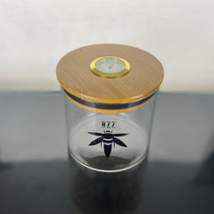 A clear glass stash jar with a bamboo lid with gold hygrometer. The glass is branded with a black Bzz Box logo.