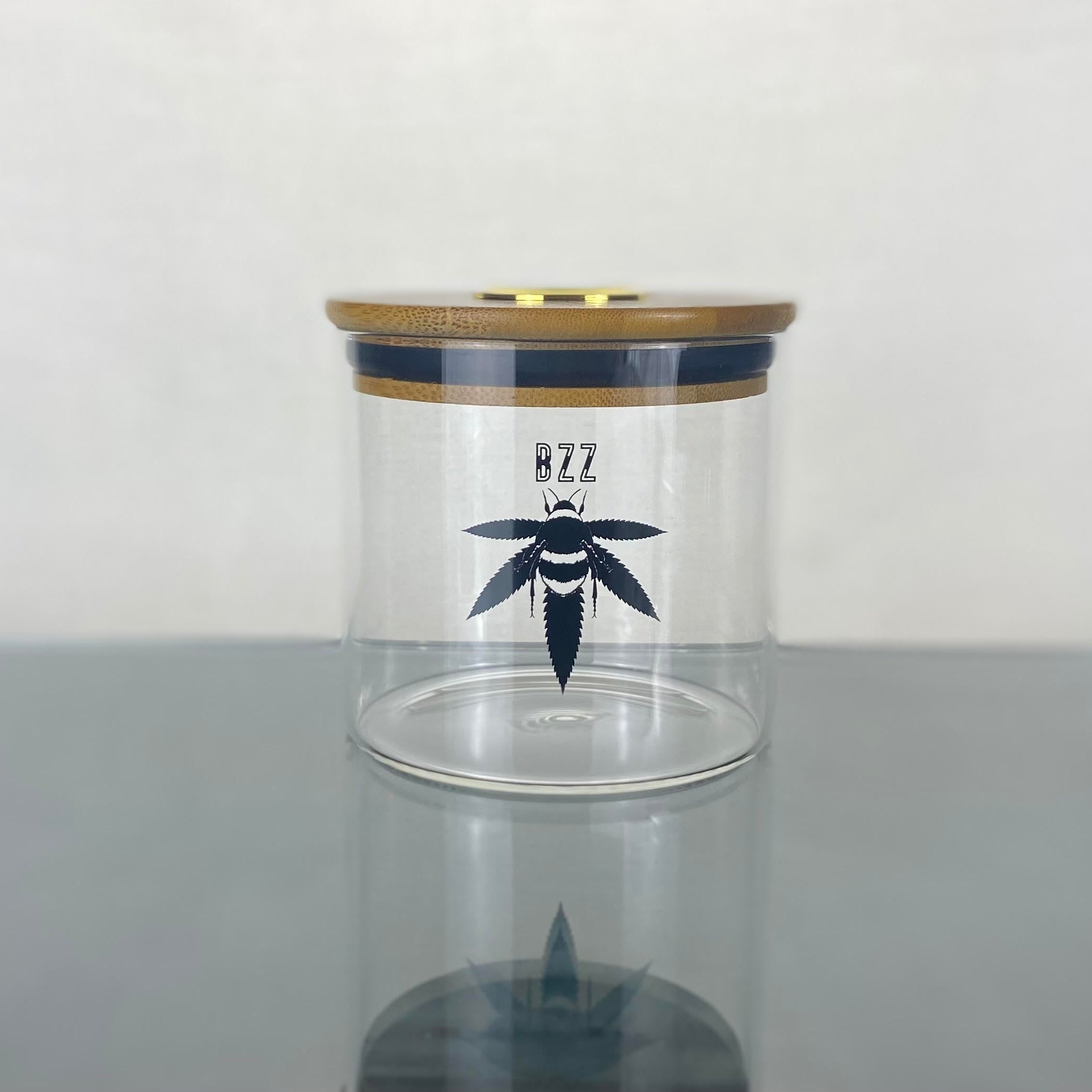 A clear glass stash jar with a bamboo lid with gold hygrometer. The glass is branded with a black Bzz Box logo.
