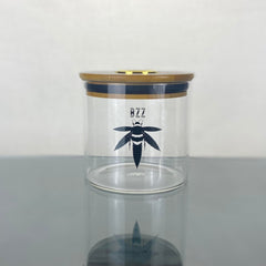 A clear glass stash jar with a bamboo lid with gold hygrometer. The glass is branded with a black Bzz Box logo.