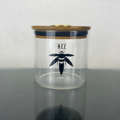 A clear glass stash jar with a bamboo lid with gold hygrometer. The glass is branded with a black Bzz Box logo.