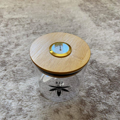 A clear glass stash jar with a bamboo lid with gold hygrometer. The glass is branded with a black Bzz Box logo.