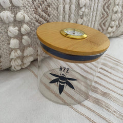 A clear glass stash jar with a bamboo lid with gold hygrometer. The glass is branded with a black Bzz Box logo.